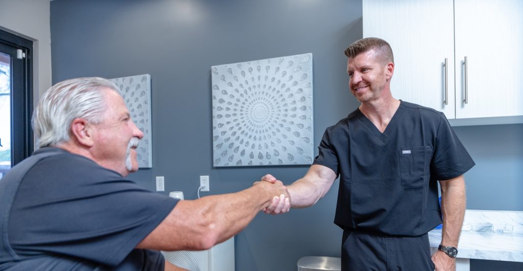 Male MedSpa provider in black scrubs shaking hands with an older male client during a consultation in a modern treatment room, conveying trust and professionalism.