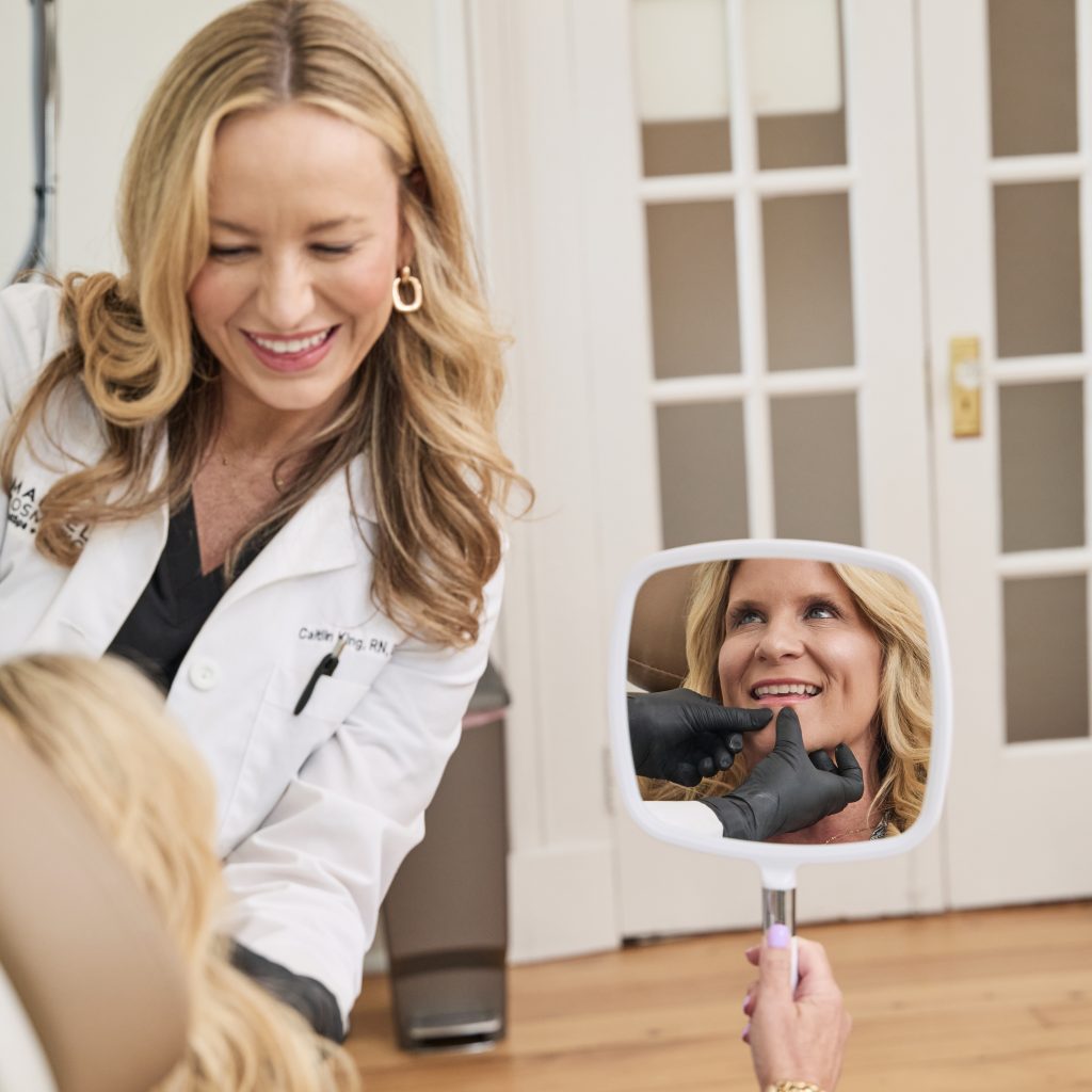 Smiling female aesthetic provider in a white coat consulting with a client holding a mirror, as the client examines her facial results during a MedSpa treatment.