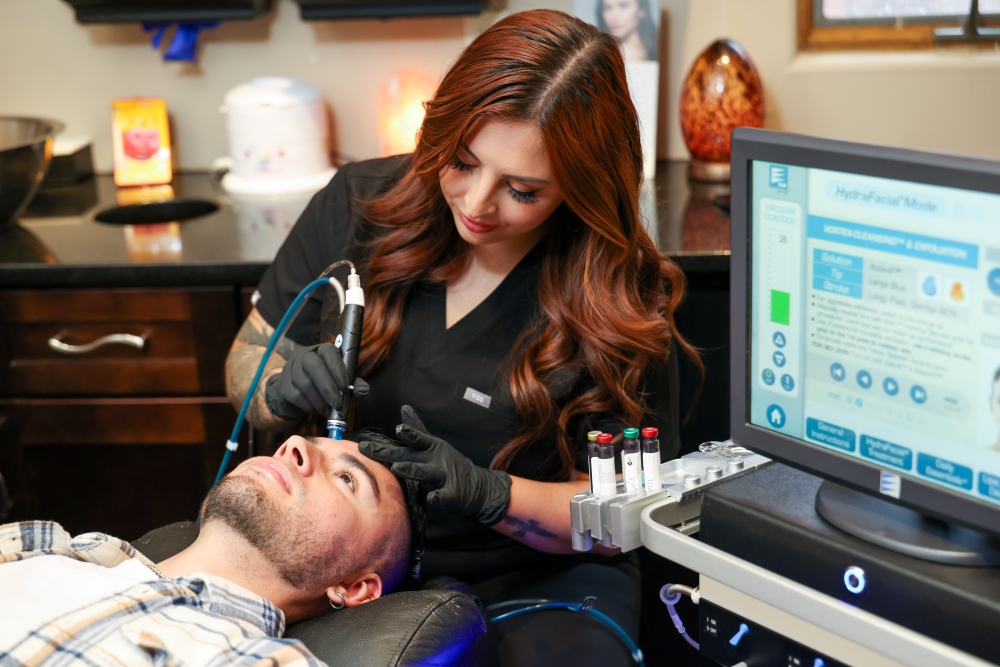 Female aesthetician administering a HydraFacial treatment to a male client using a multi-step facial device, with a digital screen showing treatment settings.