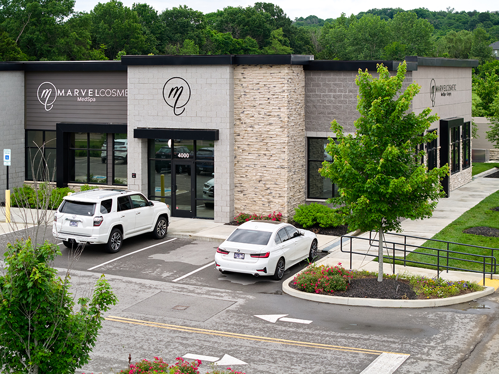 Exterior view of Marvel Cosmetic MedSpa, a modern medical aesthetics clinic with stone and gray facade, located in a suburban setting with landscaped greenery and parked white vehicles in front.