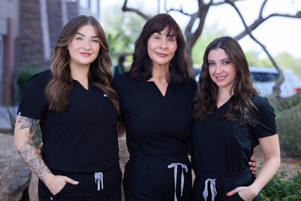Three female medspa professionals stand confidently outside a clinic, dressed in matching black scrubs, smiling and embracing a sense of team unity and professionalism.
