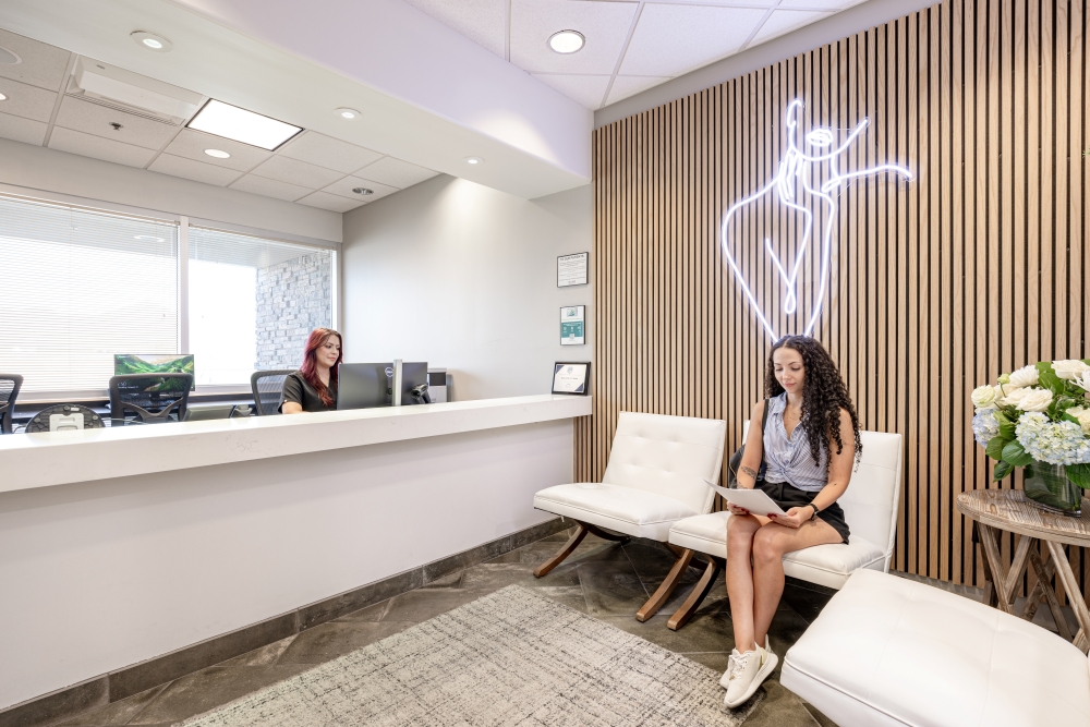 Modern MedSpa reception area with a front desk associate working and a client seated, filling out paperwork beneath a branded neon logo on a wood accent wall.
