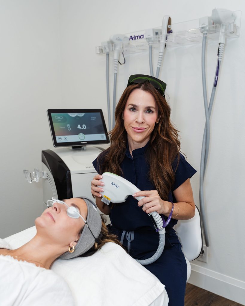 Aesthetic provider in navy scrubs prepares to perform a laser treatment using an Alma device, while a female client lies on a treatment bed wearing protective goggles in a modern med spa room.