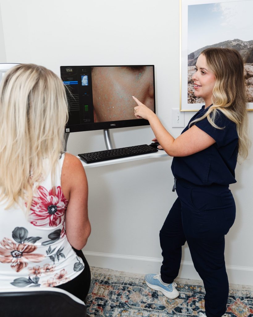 A skincare specialist in navy scrubs explains a client's facial scan results on a high-resolution VISIA screen, pointing at highlighted skin concerns while the client attentively watches.