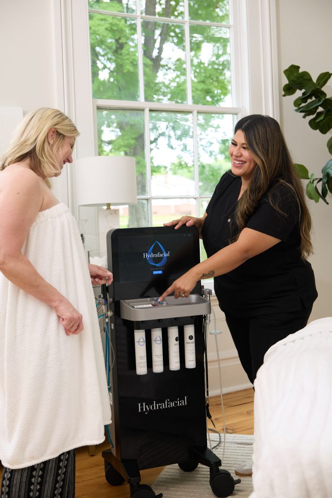 A spa technician demonstrates a HydraFacial machine to a smiling female client wrapped in a towel, standing in a bright, modern treatment room with large windows and natural light.